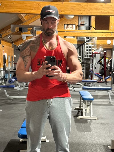 The image depicts a male subject taking a mirror selfie within a gym in Jemelle, Belgium. The mid-adult male has a muscular build, a dark beard, and mustache. He is wearing a black baseball cap with red lettering, a red tank top with white text, light grey sweatpants, a gold-colored chain necklace, and a gold-colored bracelet on his right wrist. A Polynesian-style tattoo covers his left shoulder and upper arm. Small, dark earbuds are visible in both ears. He holds a dark smartphone, likely an iPhone, at chest level with both hands, directly facing the camera. The gym floor is dark, possibly rubber matting. The walls are light-toned wood paneling, and the ceiling features exposed wooden beams. In the background, various exercise equipment, including blue-padded benches, weight machines, and a dumbbell rack, are visible. A dark spiral staircase is situated on the right side of the frame, above which a framed image of a woman's face is mounted on the wall. A circular clock is visible further to the right. The scene is lit by interior artificial lighting.