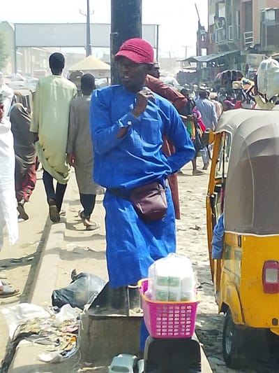 This image captures a bustling street scene in Fagge D1, Nigeria, under bright daylight. The central figure is a dark-skinned male, positioned slightly off-center to the right, wearing a blue long-sleeved tunic, dark trousers, and a red baseball cap. A dark-colored fanny pack is worn on his waist. His right hand is raised towards his mouth, while his left hand rests on his hip. He is looking towards the right side of the frame. To his immediate left (viewer's right) is the front half of a yellow auto-rickshaw with its passenger side door open. Resting on a dark, low stand or object near the auto-rickshaw is a pink plastic basket containing several small, clear plastic containers, likely products for sale, wrapped in clear plastic.
In the mid-ground on the left, several individuals are walking away from the camera on what appears to be a raised concrete curb or sidewalk. One person wears a light green long tunic, another a light brown tunic, and a partially visible individual on the far left wears white attire. The ground in the foreground on the left contains scattered debris and a black plastic bag of trash. The background reveals a dense crowd of people, various market stalls or temporary structures, and multi-story buildings lining the street, indicating a busy commercial district. The overall environment suggests an outdoor market with significant pedestrian activity and possibly some vehicular traffic. The lighting is harsh, and the atmosphere appears somewhat dusty.