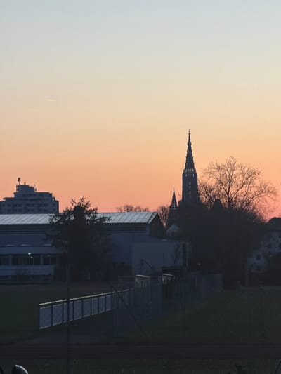 The image displays a panoramic cityscape captured from Neu-Ulm, Germany, looking towards the city of Ulm. The sky, indicating either dawn or dusk, exhibits a gradient from pale blue at the zenith to an orange-pink hue near the horizon. A faint white contrail is visible horizontally in the upper-left quadrant. The prominent Ulm Minster spire, a dark, gothic silhouette, dominates the central-right background, accompanied by a smaller, secondary spire to its left. To the far left, a multi-story building featuring multiple roof-mounted antennas is visible. The midground contains several low-rise buildings, including a large structure with a light-colored, pitched roof, partially obscured by numerous leafless deciduous trees. A chain-link fence with a horizontal metal barrier runs across the lower midground. The immediate foreground consists of a dark green field and a reddish-brown track, consistent with a sports facility. No individuals are present in the scene.