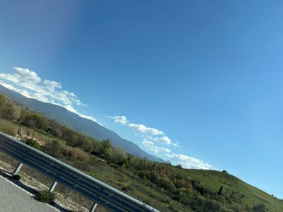 The image depicts a landscape view of Damianitsa, Bulgaria. A clear blue sky dominates most of the frame, with scattered white clouds along the horizon. The landscape transitions from rolling green hills on the left to a mountain range on the right, some parts of which are possibly snow-capped. A highway with a guardrail runs along the top of the image, adjacent to grassy vegetation. There are no discernible people or interactions visible. The environment appears rural, showcasing the natural beauty of the region.