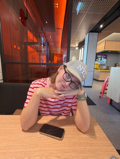 A young woman wearing a grey NYC baseball cap, black-rimmed glasses, and a pink and white striped t-shirt is seated at a wooden table in a modern fast-food restaurant. Her head is resting on her hands, which are clasped together. A smartphone lies on the table in front of her. The restaurant's interior features red-tinted glass walls, a wood-paneled ceiling with circular perforations, and brightly lit signage. In the background, a yellow service counter with a digital menu board is visible. The environment suggests a commercial establishment, possibly a McDonald's, given the visible signage. This location is identified as McDonald's Herbolzheim, Germany.