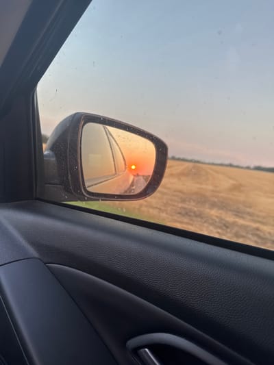The image captures a sunset viewed from inside a vehicle, looking out the passenger side window and into the side mirror. The side mirror reflects a bright orange sun low on the horizon, casting a warm glow over a harvested field of golden stubble. The field stretches towards a distant treeline under a hazy, pale blue sky. The interior of the car, including the black textured door panel and armrest, is visible in the foreground. The image is taken from a roadside perspective, suggesting a journey through a rural landscape. The location is identified as Magistralny, Russia.