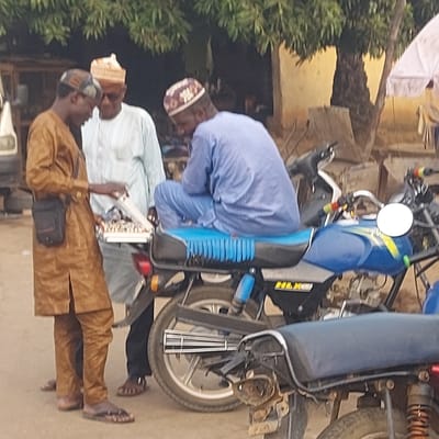 This image, presented as if rotated 90 degrees clockwise, captures an outdoor scene featuring three men and a motorcycle.
In the foreground, on the right side of the upright image, stands a man wearing a brown traditional outfit, possibly an agbada or kaftan, and a matching cap. He has a small black bag slung across his chest or shoulder. He is looking towards the left, appearing engaged with the other individuals.
In the middle of the scene, positioned slightly behind the man in brown, is another man dressed in a light-colored (possibly white or very pale blue) traditional tunic and a patterned cap. He stands near the rear of the blue motorcycle.
The third man, positioned towards the left side of the upright image, is wearing a light blue traditional tunic and a patterned cap similar to the middle man's. He is bent over, seemingly inspecting or interacting with the blue motorcycle, specifically the area around its fuel tank and handlebars. His focus is entirely on the bike.
The central object of the image is a blue and black motorcycle, which appears to be a standard commuter model. The word "HLK" is visible in white letters on a black panel on the side of the motorcycle. The seat is ribbed in blue and black. A white rectangular object, possibly a license plate, is visible near the rear wheel, but its text is illegible.
The setting appears to be a daytime outdoor environment, possibly a street, alleyway, or courtyard. In the background on the left, there is some green foliage from trees or bushes. Further in the background, out-of-focus structures or buildings are visible. A white garment is hanging on a line or structure in the upper left background. The ground beneath the individuals and the motorcycle appears to be light-colored dirt or paved surface. The lighting suggests it could be mid-day or late afternoon, with soft, diffused light. The men's attire suggests a cultural context where traditional garments are common.