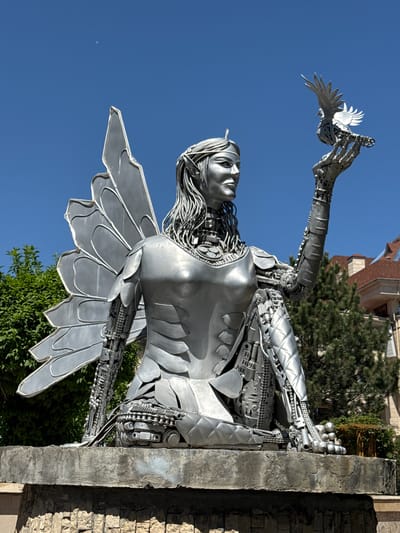 A low-angle shot shows a silver metal statue of a woman with wings, mounted on a stone base. The statue has intricate mechanical details visible in the arms, chest, and neck. The woman's left arm is extended upwards, holding a white bird. The background includes a clear blue sky, trees, and a building with a red-tiled roof. The statue appears to be located outdoors in Kenbulun, Kyrgyzstan.