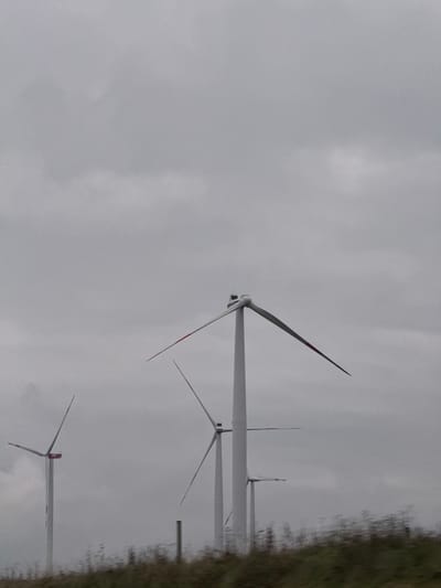 The image displays a rural landscape near Dornstadt, Germany, under an overcast sky. Four prominent wind turbines are visible, characterized by white towers and three blades each. The blades feature red tips. The turbine in the immediate right foreground has its blades oriented in an upward V-shape. Other turbines are situated further in the background. A smaller, partially obscured wind turbine is discernible in the far background to the right. The foreground shows a blurry strip of green and brown vegetation, consistent with a field or hillside, with a single dark vertical post positioned to the left of center. The sky is uniformly light grey and overcast, lacking distinct cloud formations. No individuals, animals, or discernible actions or interactions are present in the frame.