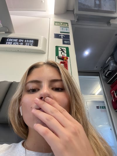The image depicts the interior of a train, contextually located in Tres Cantos, Spain. A young light-skinned female with blonde hair occupies the foreground, looking directly towards the camera, her right hand partially covering her mouth. She wears a white earbud in her left ear. In the upper left quadrant, a digital display indicates "COCHE 04 13:56 190 KM/H 31°C". To the right of the display, various train interior signs are visible, including green and blue pictograms for "Exit", "WC", and directional arrows. A red emergency hammer device is mounted below the emergency exit sign. Overhead luggage compartments containing gray and red bags are visible in the upper right, and a grey seat is partially visible on the left.