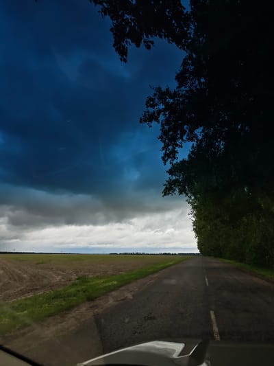 The image depicts a road traveling through the countryside near Zhornyshche, Ukraine. The road stretches into the distance between a cultivated field to the left and dense foliage on the right. The sky is dominated by dark, ominous clouds, indicating an approaching storm. The vantage point is from inside a vehicle, with the car's hood partially visible at the bottom of the frame. There are no people visible. Electrical power lines are visible on the distant horizon.
