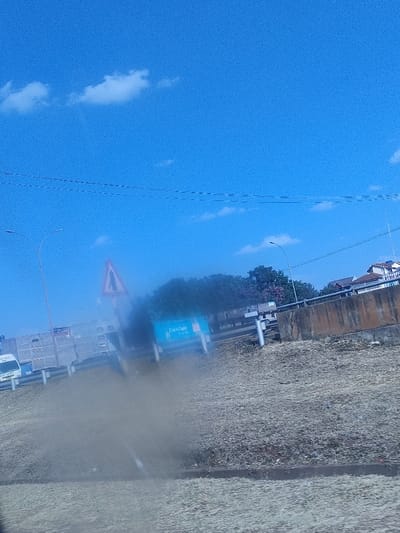 A hazy view from inside a vehicle shows a highway scene on a sunny day. The blue sky has scattered clouds and overhead electrical wires. A road sign and a billboard are partially obscured by the hazy view. A white van and some buildings are visible in the background, along with a concrete barrier and roadside grass. The haze mostly obscures the lower half of the image.