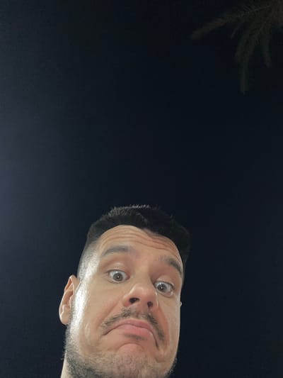 A close-up, low-angle, outdoor night photograph shows the upper portion of a man's face and his head. He is looking up at the camera with wide, surprised eyes and a pouting mouth. His hair is dark and cut short on the sides, with longer, spiky hair on top. He has a mustache and stubble on his chin. His forehead is wrinkled, and his skin appears slightly sweaty. The background is a dark, almost black sky, with the faint silhouette of a palm frond visible in the upper right corner. The man's head and shoulders fill the bottom half of the frame, with the sky occupying the top half. The image appears to have been taken in Roquetas de Mar, Spain, possibly near a park or beach area where palm trees are common.