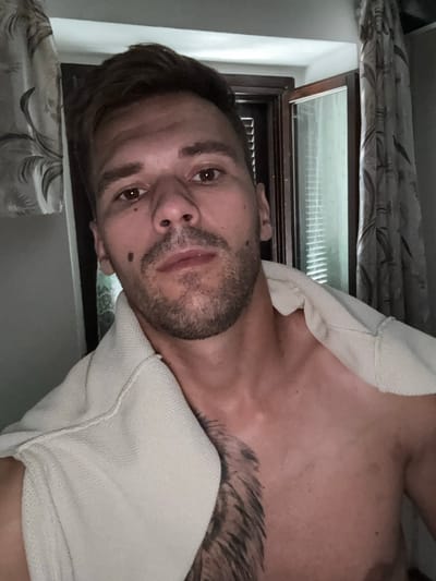 The media file is a close-up, rotated photograph of a person. The subject is a man with short, dark hair and stubble. He has brown eyes and a mole on his left cheek. He appears to be wearing a light-colored garment, possibly a t-shirt, draped around his shoulders. A tattoo is visible on his upper chest. The background shows a window with blinds and curtains, suggesting an indoor setting. The lighting is dim, indicating it might be evening or nighttime. No text is visible in the image. The overall mood is relaxed and informal. The location context is Sesto Calende, Italy, though no specific visual cues in the image confirm this.