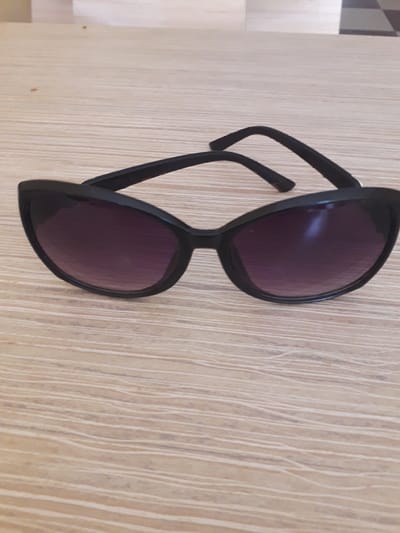 The image displays a pair of black-framed sunglasses resting on a light-colored wooden surface. The sunglasses are positioned diagonally across the frame, with the lenses and main body in the central and lower right, and one arm extending towards the upper left. The frames are solid black, with a slightly rounded, possibly subtle cat-eye style. The lenses are dark purple or grey-tinted and show minor reflections of ambient light. The temples (arms) of the sunglasses are folded.

The background consists of a wooden surface, characterized by a light, pale brown or beige color with prominent, fine vertical grain lines that are lighter in tone, creating a textured appearance. A blurred area in the far left background suggests a corner or edge of another surface. No activity is taking place; the sunglasses are stationary. There are no people visible. The time of day, weather, or specific location cues beyond the wooden surface are not discernible. No text is visible on any object in the image. This scene is set within the general location context of Ghafurov, Tajikistan.