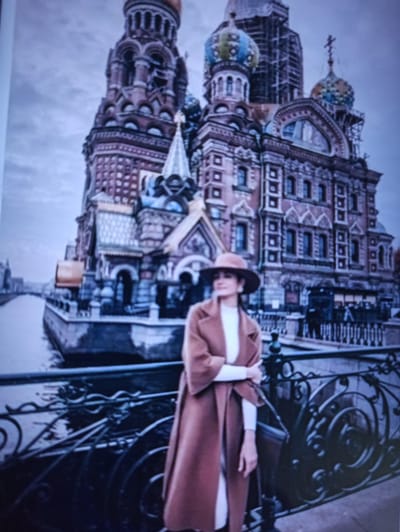 A person wearing a brown hat and coat stands on a bridge in front of a large, ornate building with multiple domes and spires, resembling the Church of the Savior on Spilled Blood in St. Petersburg, Russia. The building is constructed of red brick with intricate decorative elements. A canal or river runs alongside the building. The sky is overcast and the overall atmosphere is muted. The person is facing to the left, with their left hand resting on the bridge railing. The image is a photograph of a photograph, with some grain and blurriness. There is no indication that the location is Balakliya, Ukraine.