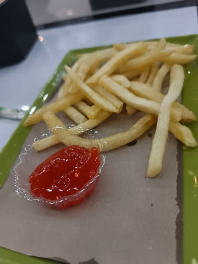 The image displays an overhead, slightly angled view of food items served indoors in Namplohbaro, Indonesia. A rectangular green plastic tray holds a light brown paper liner, upon which a scattered pile of golden-brown french fries is present. A transparent, fluted plastic cup containing a vibrant red sauce, consistent with ketchup, is positioned in the lower-left portion of the liner. The french fries exhibit typical stick shapes with varying lengths and slight surface imperfections. No individuals are visible. The background consists of blurred light and dark surfaces, typical of an interior setting.