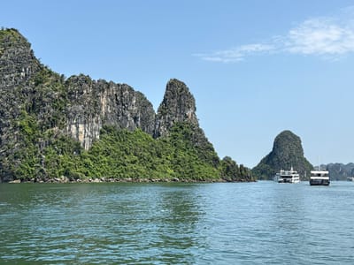 This image captures a scenic daytime view of Hạ Long Bay, near Tuan Chau Ward, Vietnam. The foreground features calm, greenish-blue water, hinting at gentle ripples. On the left and extending across the mid-ground, majestic limestone karsts rise dramatically from the bay. These towering islands are heavily adorned with dense green vegetation, contrasting with the grey, rugged rock faces.

The clear, bright blue sky with a few wispy white clouds suggests a sunny day, likely late morning or early afternoon. In the mid-ground on the right, two multi-deck boats are visible. The closer boat has a white superstructure and a dark hull, seen from its stern. Behind it, a larger, predominantly white cruise ship with dark windows is navigating through the water, with another verdant karst island in the background. The presence of these vessels indicates active tourism or sightseeing in the bay. No visible text is present in the scene. The overall impression is one of natural beauty and tranquility.