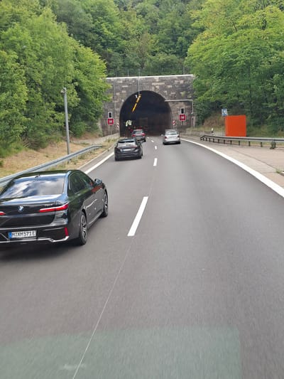A black BMW 7 Series sedan with license plate "M X M 571 E" is captured from a low-angle perspective, driving on a multi-lane highway towards a tunnel entrance in Wiesensteig, Germany. The car is positioned in the foreground, on the right lane of the highway. Other vehicles, including a silver hatchback and another dark sedan, are visible ahead, approaching the tunnel. Red speed limit signs indicating "100" are mounted on poles to the left of the tunnel entrance. The tunnel is constructed with stone and has lights illuminating its interior. Lush green trees and vegetation surround the highway and the tunnel. The sky is overcast, and the road surface appears to be wet in places.