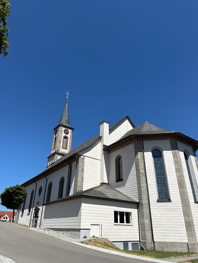 The image shows the Saint Michael's Catholic Church in Schönwald im Schwarzwald, Germany, on a clear, sunny day. The church is a white building with a prominent steeple that features a clock and a cross at its apex. The exterior walls are a combination of white siding and grey stone accents. Arched windows with stained glass are visible on the rounded section of the church. A small, green tree stands to the left of the building, and a paved road slopes upwards towards the church in the foreground. The sky is a vibrant blue with no clouds.