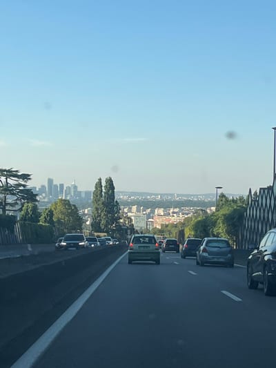 The image depicts a view from a highway, likely a French Autoroute, with a distant cityscape in the background. The foreground is dominated by a multi-lane road with several cars driving. The sky is a clear, light blue. To the left, there's a concrete barrier and some greenery, including trees and bushes. In the mid-ground, the landscape transitions to a more urban setting with buildings and more trees.
The prominent feature in the background is a cluster of tall buildings, characteristic of a business district, appearing to be La Défense in Paris. The overall scene suggests a sunny day, possibly late afternoon, given the lighting. The composition is from the perspective of someone within a vehicle, looking forward and slightly to the left.
The city of Sèvres is known to be located southwest of Paris, and the view of the La Défense skyline from an elevated position, such as a highway overlooking the city, is consistent with this geographic context. The presence of traffic and the highway infrastructure further supports this interpretation.