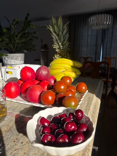 The image depicts a collection of fresh fruit arranged on a kitchen countertop. In the foreground, a white, scalloped bowl contains a pile of ripe cherries. To the left, a glass with a golden liquid, likely whiskey, sits on the counter. A white tray holds a variety of red plums or nectarines, some with a slightly purplish hue. Interspersed with these are small, dark red tomatoes, some showing hints of green. In the background, a bunch of bright yellow bananas is partially visible. Behind the fruit, a tall pineapple stands upright. Further back, a green leafy plant in a white textured pot is illuminated by sunlight streaming in from the right. The environment suggests an indoor setting, possibly a home kitchen, with elements like a chandelier and dining chairs visible in the dimly lit background. The presence of a modern chandelier and the overall style of the furniture might suggest a contemporary residential interior. There are no people in the image. The image does not provide any information to associate the location with Dieppe, Canada.