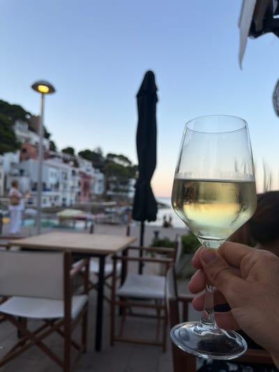 The image shows a person holding a glass of white wine in a coastal town in Begur, Spain. The background is blurred, but it appears to be an outdoor seating area of a restaurant or bar, with tables and chairs visible. Beyond that, there are buildings with white and pink facades cascading down a hillside towards the sea. The sky is a clear, pale blue, suggesting it is late afternoon or early evening. A street lamp is illuminated on the left side of the image. A folded black umbrella is also visible behind the glass. The wine in the glass is a pale yellow color. The person's hand, holding the stem of the glass, is in focus in the foreground. The base of the wine glass has a visible etching of "VICRILA."
