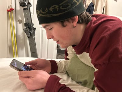 A young person with a dark beanie and a maroon and beige striped shirt lies in bed using a smartphone. They have short brown hair and a silver hoop earring. Behind them, skis and poles lean against a white door. The room appears to be a bedroom, with bedding visible. The person is looking intently at the phone screen, their hands holding the device in landscape orientation.