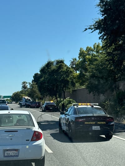 The image shows a traffic incident on a sunny day in North Highlands, United States. A white Chevrolet Cobalt is in the foreground, facing away from the camera, with its license plate visible as "7AHJ708". To the right and further down the road, a black Dodge Charger with "HIGHWAY PATROL" on the back and license plate "15L8990" is parked. A tow truck is visible in the middle of the road, assisting a red car. Several other vehicles and individuals, including what appear to be law enforcement officers, are present at the scene. The incident is taking place on a multi-lane road surrounded by trees and vegetation, with a clear blue sky overhead.