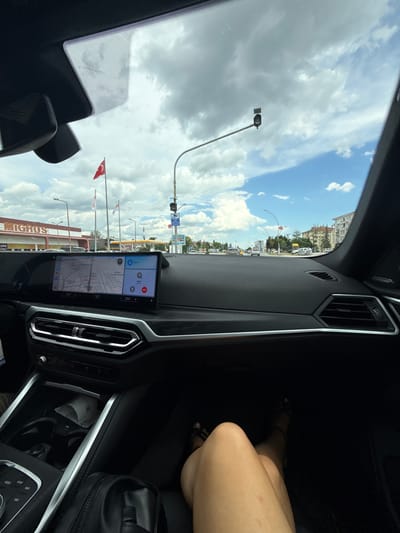 The image, taken from inside a car, shows a view of Edirne Merkez, Turkey. Through the windshield, the sky is visible with a mix of blue and cloudy areas. A traffic light, street lights, and Turkish flags are seen along the road. A building with the name "MIGROS" is also visible. The car's dashboard features a navigation screen displaying a map and controls. Part of the passenger seat is visible, showing a person's leg and a black bag.