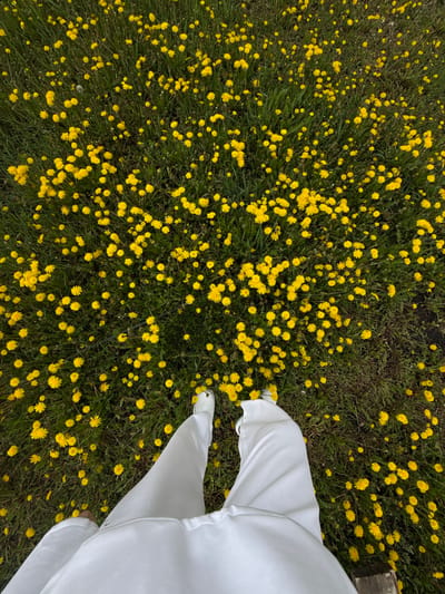 An aerial perspective shows a grassy field densely populated with blooming yellow dandelions. In the lower right corner of the image stand a person wearing white pants and white perforated shoes with visible silver lace tips. The scene is in an outdoor setting. The location, if Ленино, Russia, is not definitively verifiable from the image itself.