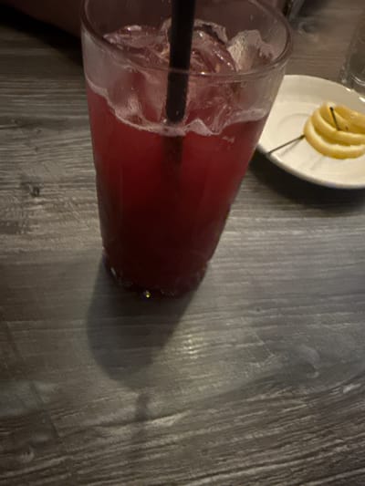 A close-up, overhead shot depicts a glass of red liquid, likely a beverage, filled with ice and a black straw. The glass sits on a gray, textured surface, possibly a wooden table. A small white plate with several lemon slices is partially visible in the upper left corner of the image. The location in Glenview, United States of America, cannot be determined from the image alone.