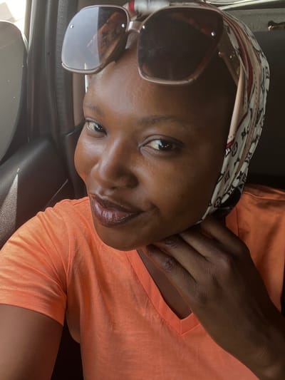 A forensic examination of the image reveals a close-up portrait of an adult female located within the interior of a vehicle in Kunkwaba, Nigeria. The subject, exhibiting dark skin and dark eyes, is positioned facing forward and slightly to the viewer's right, directly engaging the camera with a subtle smile. Her lips are accented with a glossy, reddish-brown cosmetic. She is attired in a bright orange V-neck t-shirt. A light-colored headscarf with a repeating pattern, consistent with a monogram or brand logo, covers her hair. Brown-framed sunglasses with dark lenses are perched on her forehead, resting atop the headscarf. Her right hand is visible, with fingers gently curled and positioned near the chin and lower jawline. The background shows elements of a vehicle's interior, including a dark panel and a partially visible window on the left, indicating natural light ingress from the exterior.