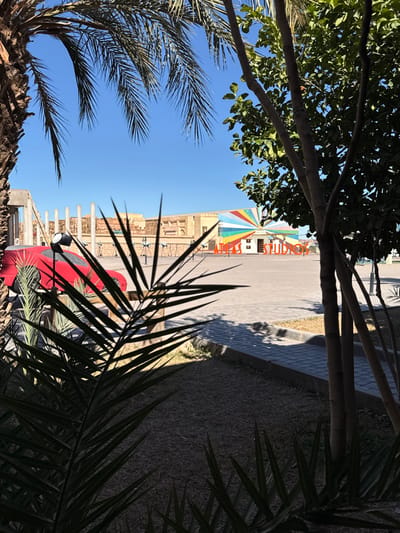 The image displays a sunlit outdoor scene at Atlas Studios in Ouarzazate, Morocco, under a clear blue sky. In the foreground, dark green palm fronds and the trunk of a large palm tree obscure the left side, while multiple slender tree trunks with green leaves occupy the right, casting shadows onto the sparse, dry ground. A paved walkway separates this vegetation from a large, unoccupied paved plaza in the midground.
On the left side of the plaza, a partially covered red sports car is positioned next to a black studio light on a stand. Numerous small, dark, tripod-like stands are scattered across the plaza. The background features light brown, sand-colored architectural facades, resembling ancient or desert structures. Centrally located in the background is a building adorned with a vibrant, multi-colored sunburst or rainbow mural. The words "ATLAS STUDIOS" are visibly inscribed in orange lettering on the ground directly in front of this building. No individuals are present, and no human activity is depicted.