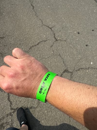 This close-up, high-angle shot captures a person's right forearm and hand, wearing a bright green wristband, against a cracked asphalt background. The arm is hairy, suggesting an adult male.
The most prominent object is the neon green wristband, which bears the text "BATA AGRO" in black capital letters. Between "BATA" and "AGRO," there is a clear black logo depicting the silhouette of a tractor, indicating an agricultural theme. This wristband likely serves as an admission pass or identification for an event.
The setting is outdoors, on a textured, dark grey asphalt pavement showing several fine cracks, suggesting age and wear. The strong shadows, including a partial shadow of a person's leg and a black shoe in the lower right corner, indicate bright sunshine and a daytime setting.
The image suggests the subject is attending an agricultural exhibition or fair, specifically "BATA AGRO," likely held in Madzherito, Bulgaria, given the provided location context. No particular activity beyond attendance is visible, and no emotions are discernible from the partial view of the person.
