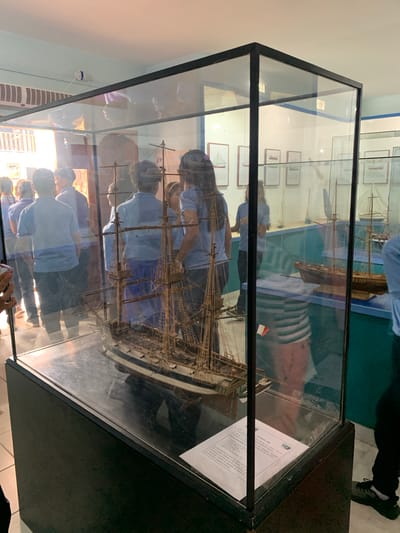 The image shows an indoor exhibition, possibly a museum, in Boca del Río, Venezuela. The main subject is a detailed scale model of a tall ship displayed in a large glass case with a black base. Several people, appearing to be visitors or students, are gathered around the display case, looking at the model. They are mostly wearing light blue shirts. In the background, other display cases containing smaller ship models and framed pictures of sailing vessels are visible. The lighting suggests an artificial indoor environment, possibly with natural light filtering in from the left side. The overall scene depicts an educational or cultural exhibition focused on maritime history.
