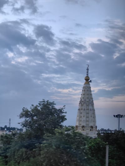 The image shows a white, tiered temple spire rising against a cloudy, pale blue sky. The spire is decorated with horizontal bands and has a golden finial topped with a small antenna. In the foreground, lush green trees obscure the lower portion of the temple and the surrounding landscape, which includes hints of buildings and a tall metal tower in the distance. The overall lighting suggests it is either early morning or late evening. This scene is located in Uchana, India.