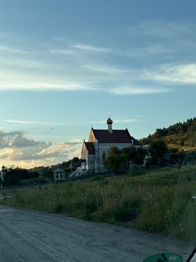 The image depicts a view of a church and its surroundings, likely in the city of Rohachyn, Ukraine, based on the provided information. The church, with a red-tiled roof and a golden dome topped with a cross, is situated on a grassy hillside. Adjacent to the church, there is a smaller, white gazebo-like structure with a dome. The foreground shows a dirt road, and the background consists of trees and hills, with a cloudy sky above. The lighting suggests it is late afternoon or early evening, casting long shadows and illuminating the clouds with warm tones. There are no people visible in the image. The overall scene is serene and rural.