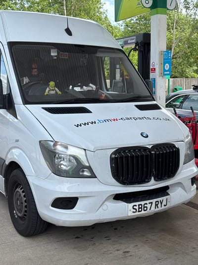 This media file shows a white van with a custom BMW front grill parked at a petrol station. The van has the website "www.bmw-carparts.co.uk" printed vertically on its side. A license plate that reads "SB67 RYJ" is visible on the front of the van. The petrol station has a visible pump with "diesel" indicated, and signage for "BP" and "No smoking." A small figure of Shrek is visible inside the van. The scene is outdoors, with trees in the background, suggesting daylight. The overall impression is a functional vehicle at a routine stop.