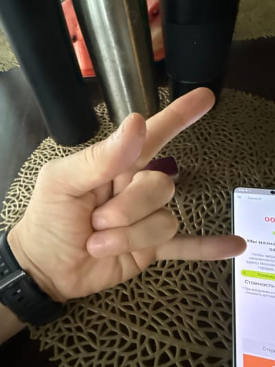 A hand with fingers extended in a "rock on" gesture is held in the foreground. The hand is positioned over a dark brown, textured placemat. To the left of the hand are dark cylindrical objects, possibly drink tumblers. To the right, a smartphone screen is visible, displaying Russian text and financial data. The text on the screen mentions "Биржа" (Stock Exchange) and details about delivery to Moscow, including cost and a notification about potentially higher delivery fees. There is no visible indication that the scene is located in Lesnoi Gorodok, Russia.