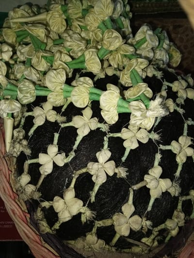In Na Sang, Laos, a dark, densely textured spherical object, resembling a large hair bun or wig composed of black fibers, is displayed within a woven basket. The object is elaborately adorned with two distinct patterns of light yellow fabric flowers. The upper portion features numerous tightly packed, bulbous flowers, each with a short green fabric stem, forming a voluminous garland that cascades over the top. Below this, across the surface of the dark sphere, smaller, flatter, four-petal-like fabric flowers are arranged in a lattice pattern, connected by thin, light-colored threads or delicate fabric stems, some showing frayed ends. The spherical item rests on and against the coarse, interwoven strands of a light brown, reddish-brown, and dark brown natural fiber basket or container, visible along the bottom and left periphery. A partial red label with indistinct text is faintly visible beneath the basket's edge on the lower left.