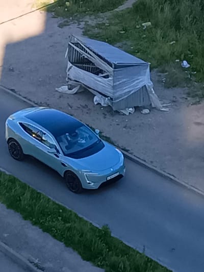 The image depicts a light blue electric car with a black roof, driving on a paved road in Kokhma, Russia. The car appears to have a modern, coupe-like design. The driver and a passenger are visible inside the vehicle. To the right of the road, there is a dirt area with some scattered debris and a makeshift shelter constructed from corrugated metal and plastic sheeting, which appears weathered and damaged. A strip of green grass runs along the left side of the road. The overall scene suggests an urban or semi-urban environment, possibly an industrial or less-developed area, given the presence of the shelter and debris.