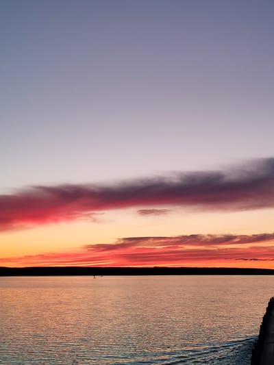 The image depicts a sunset over a body of water, likely a lake or large river. The sky transitions from a pale lavender at the top to vibrant oranges and reds near the horizon. A dark band of trees is visible on the horizon line. The water reflects the colors of the sky. There are two figures standing or floating on the water. The location is unspecified but may be Topornya, Russia.