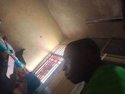 The image depicts an indoor scene in Rumuekini, Nigeria, featuring two individuals and elements of a room. On the right side of the frame, a person with dark skin and short hair is seen in profile, looking towards the left. They are wearing a bright green t-shirt, and their expression appears neutral.
On the left, another person's hand is visible holding a smartphone, with its screen illuminated. A colorful, patterned fabric, possibly a blanket or clothing in shades of blue, pink, and yellow, is also visible next to the hand.
The room's ceiling, a light beige or off-white color, shows some discoloration and textures. A fluorescent light fixture is mounted on the ceiling towards the upper right. Natural light enters the room from a window on the right side, which is characterized by vertical bars, suggesting security features or a specific architectural style. The overall lighting in the room is dim, with the window being the brightest light source.
The activity taking place involves one person observing, potentially looking at the smartphone held by the other individual, who is actively interacting with the device. The time of day appears to be daytime due to the natural light. There is no visible text in the image.