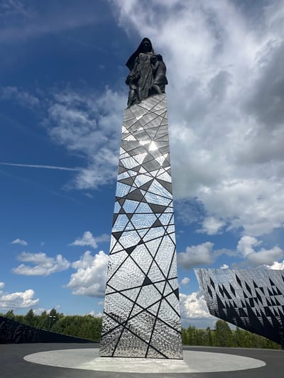 A tall, slender monument with a dark, silhouetted sculpture of a hooded figure holding two children stands at the top. The monument's shaft is clad in reflective, textured panels arranged in a geometric pattern of interconnected triangles and quadrilaterals. The background features a bright blue sky with scattered white clouds, and in the distance, a dense green forest is visible. To the right of the main monument, another angular, reflective structure is partially visible. The scene appears to be outdoors, possibly in a memorial park or public space in the city of Izhora (Verevskoe S/P), Russia. The ground in the foreground is paved with a dark, curved pathway and a lighter circular area.