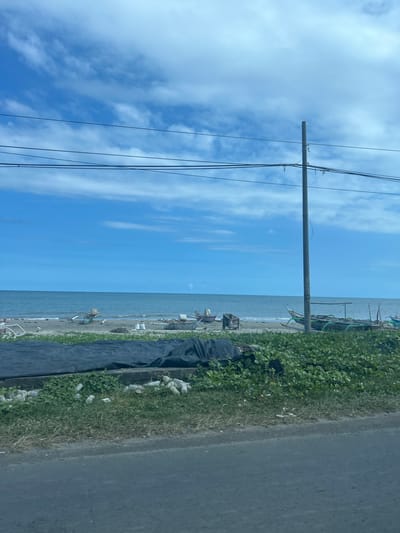 The image depicts a coastal scene in Mariorennu, Indonesia, featuring a body of water, a beach, and a road. In the foreground, a paved road runs horizontally across the bottom of the frame. Adjacent to the road, a strip of green vegetation is visible, interspersed with what appear to be white cylindrical objects. Further back, a large black tarpaulin covers an area of ground. Beyond the tarpaulin, the sandy beach is lined with several small fishing boats of varying sizes, some partially obscured by vegetation. The sea extends to the horizon, meeting a bright blue sky dotted with scattered white clouds. A utility pole stands tall on the right side of the image, with wires strung across the sky. The overall impression is one of a somewhat undeveloped coastal area, likely used for fishing activities.
