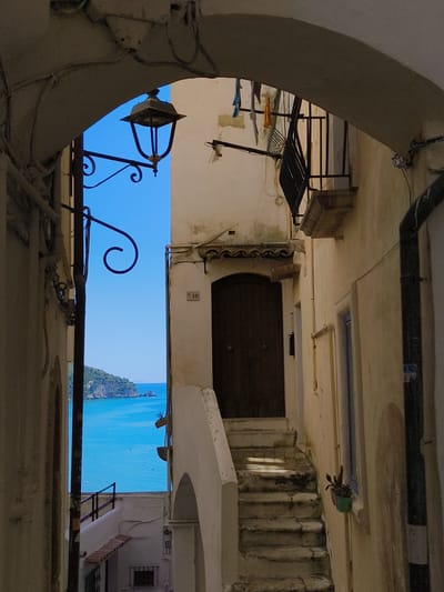 This is a photograph of a narrow alleyway in Sperlonga, Italy, looking out towards the sea. The alley is framed by the arches of buildings on either side, and a wrought-iron lamppost is visible on the left. A set of outdoor stairs leads up to a wooden door on the right, with a small balcony above it. Laundry is hanging on a line across the alleyway. The view through the alley opens up to a bright blue sea and a distant coastline with green hills. The buildings are made of stucco and are painted in a light, neutral color. The sun is casting shadows on the stairs, suggesting it is daytime. The overall impression is of a charming, sun-drenched Mediterranean street.