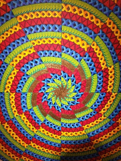 A vibrant, radial pattern dominates the scene, featuring concentric bands of geometric shapes. Triangles, diamonds, and x-shaped patterns repeat in blocks of red, blue, yellow, and green, creating a visual swirl. The center features a starburst design in metallic tones, surrounded by segments of brown and blue. The overall effect is a dynamic and colorful geometric composition with a focal point at the center.