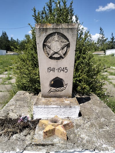 The image shows a stone monument in Cinișeuți, Moldova, commemorating the years 1941-1945. The monument features a prominent star carving at the top, with the dates inscribed below. A relief of a helmet and laurel wreath is visible beneath the dates. The monument is surrounded by greenery, with a clear blue sky and scattered clouds in the background. In the foreground, there are weathered stone slabs and a smaller, star-shaped structure, possibly a memorial or offering area, which appears to be made of bricks and is partially damaged. A pink plastic bag with some writing on it is visible on the ground near the base of the monument. The overall scene suggests a memorial site, possibly a war memorial, located outdoors.