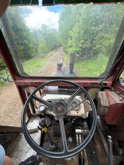 The image is an interior view from the driver's seat of a vehicle, possibly a tractor or a similar type of machinery. The environment outside the vehicle shows a rural area with a dirt road or path surrounded by trees and vegetation. The cabin of the vehicle is rudimentary, with a large steering wheel dominating the foreground. Various gauges and levers are visible on the dashboard. The vehicle appears to be located in a wooded area in the city of Рилски манастир, Bulgaria.