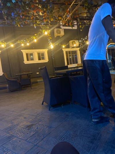 The image displays an outdoor or semi-enclosed patio area in Igbessa, Nigeria, characterized by an evening ambiance. The ground is paved with dark, stamped concrete exhibiting patterns resembling wooden planks and decorative textures. A dark-colored building wall forms the background, featuring two white-trimmed windows and two white air conditioning units mounted above them. Overhead, numerous string lights with spherical bulbs are suspended and interwoven with artificial green and multi-colored foliage, serving as the primary light source.
Furniture includes several dark, possibly wicker or rattan, seating arrangements. In the midground, a square table with a glass top is positioned with two matching chairs. To the left, a smaller round table is accompanied by two similar chairs.
One individual is visible on the right side of the frame, from the mid-torso down, wearing a white short-sleeved t-shirt, dark pants, and dark open-backed footwear. This person appears to be standing or in motion. A second individual's head and shoulders are dimly visible in the distant background, positioned behind the midground furniture. No direct interactions between individuals are discernible.