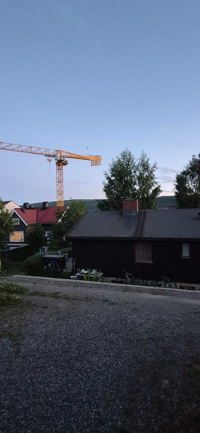 The image depicts a construction crane in a semi-urban or rural setting, possibly in or near Lillehammer, Norway, given the context. The sky is clear and blue, suggesting daytime, though the lighting is subdued, hinting at early morning or late evening.

In the background, a large yellow construction crane dominates the skyline. Its boom extends horizontally from a central tower. The crane appears to be on a construction site, indicated by its presence amongst buildings.

In the middle ground, several residential buildings are visible. One building has a dark facade and a dark-colored roof, with a brick chimney. Adjacent to it is another building with a bright red roof and dark walls, featuring a lit window. Trees are scattered throughout this area, some with dense green foliage.

In the foreground, a gravel-covered area or driveway leads up to the buildings. Several bicycles are parked near the dark-colored building in the middle ground. There is also some scattered debris or construction materials on a table or platform.

The overall impression is one of a quiet, possibly residential area with ongoing construction work. The scene is static, with no human presence immediately apparent. The environment appears to be somewhat unkempt, with vegetation growing around the buildings and debris in the foreground.