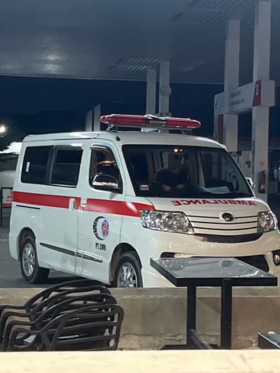 The image shows a white ambulance parked next to an outdoor seating area. The ambulance has red stripes and the word "AMBULANCE" written in red letters on the front. The side of the ambulance has the words "PT. GMN" and an emblem. The location appears to be a public space or business in Meunasahceubrek, Indonesia, as inferred from the emblem on the ambulance. The seating area has multiple black metal chairs. The surrounding environment seems to be illuminated by artificial lighting, suggesting it is night.
