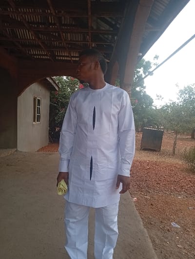 An adult male stands on a light-colored concrete path in Ugwolawo, Nigeria. He is dressed in a white long-sleeved tunic with two dark vertical panels on the front and matching white trousers, holding a rolled yellow cloth in his left hand. His head is turned left in profile, with his gaze directed slightly upwards. Overhead, a wooden beam structure with corrugated metal roofing and arched supports is visible. To the left of the subject, a light-colored building with a single white-framed window and red flowering bushes is partially visible. The right background consists of reddish-brown earth with sparse vegetation and a dark, rectangular container. The scene is illuminated by natural daylight.